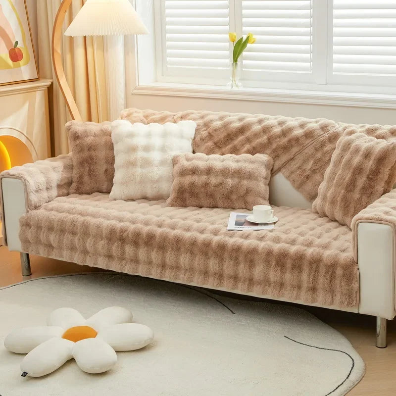 Housse de canapé en fausse fourrure de couleur beige avec des coussins en gradation de beige à blanc, créant une ambiance cosy et élégante dans un salon éclairé naturellement